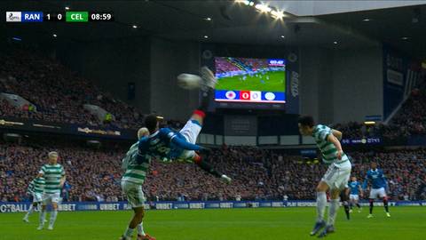 Piłkarz wykonuje akrobatyczną przewrotkę, pr&oacute;bując strzelić gola. Wok&oacute;ł niego znajdują się inni zawodnicy i liczni kibice na trybunach stadionu.