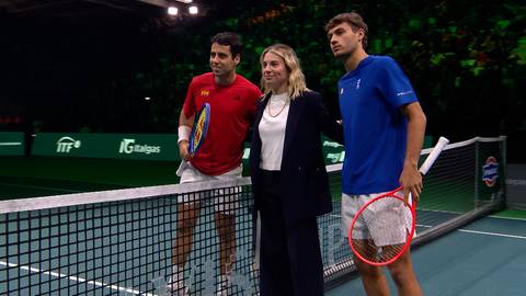 Dwóch mężczyzn i kobieta stoją na korcie tenisowym. Mężczyzna po lewej ma czerwoną koszulkę, a po prawej niebieską. Kobieta pośrodku ubrana jest w ciemny żakiet.