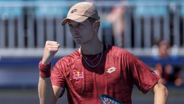 Tenisista w czerwonej koszulce i czapce z daszkiem, z zaciśniętą pięścią i uśmiechem na twarzy, trzyma rakietę tenisową.