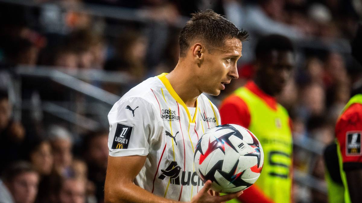 Piłkarz w białej koszulce z żółtym kołnierzykiem trzyma piłkę nożną w rękach.