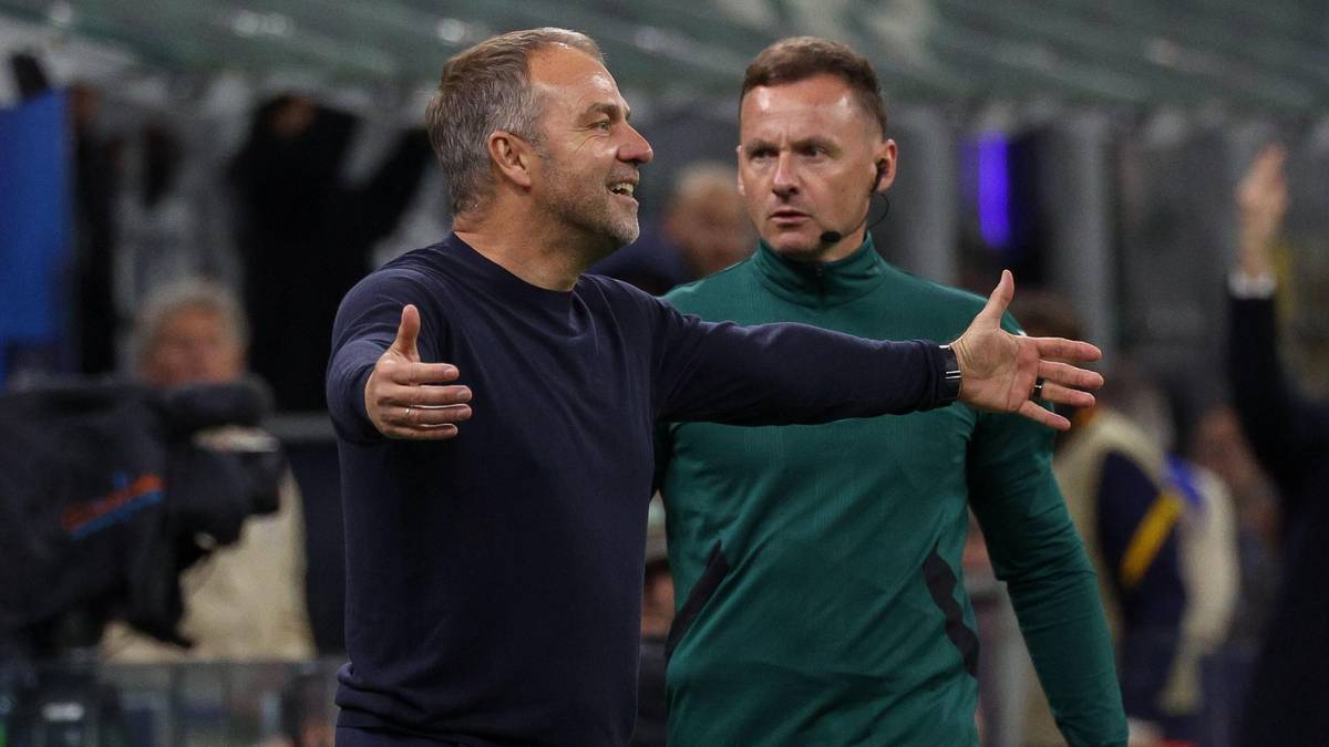 Hansi Flick, w granatowej bluzie, z podniesionymi kciukami i rozłożonymi ramionami, rozmawia z innym mężczyzną w zielonej bluzie.