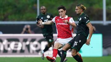 Dw&oacute;ch piłkarzy w akcji na boisku piłkarskim, jeden w czerwonej koszulce, drugi w czarnej, walczący o piłkę.