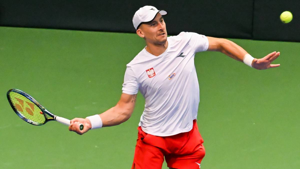 Polski tenisista w białej koszulce z godłem Polski i czerwonymi spodenkami, podczas meczu na korcie. Uderza piłkę rakietą tenisową.