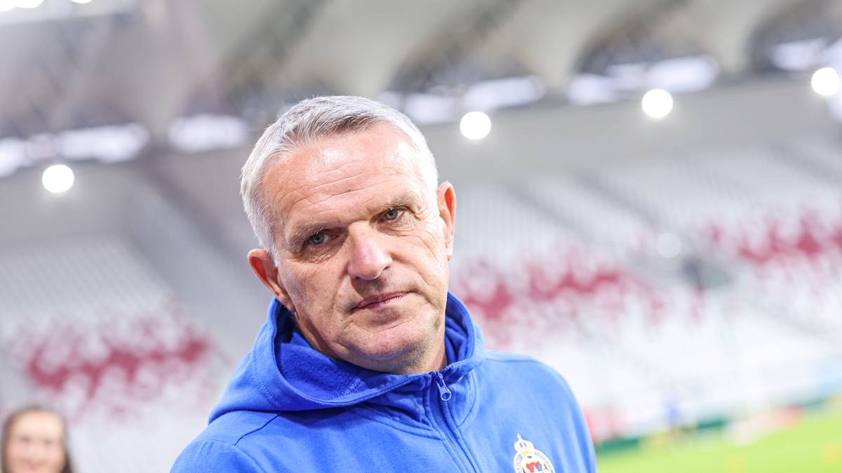 Portret Kazimierza Moskala, trenera piłkarskiego, w niebieskiej bluzie na tle trybun stadionu.