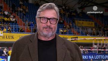 Wojciech Drzyzga, ekspert siatkarski, w okularach i brązowej marynarce, na tle trybun hali sportowej.