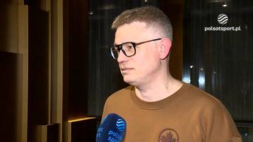 Przemysław Pawlak w okularach w brązowej bluzie z logo, udziela wywiadu przed ciemnym tłem z pionowymi elementami.