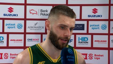 Portret koszykarza z brodą, udzielającego wywiadu przed ścianą z logotypami sponsor&oacute;w.