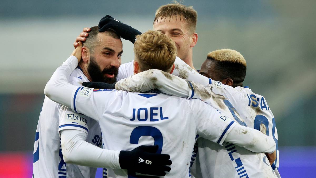 Pięciu piłkarzy w białych strojach celebruje bramkę, przytulając się do siebie.