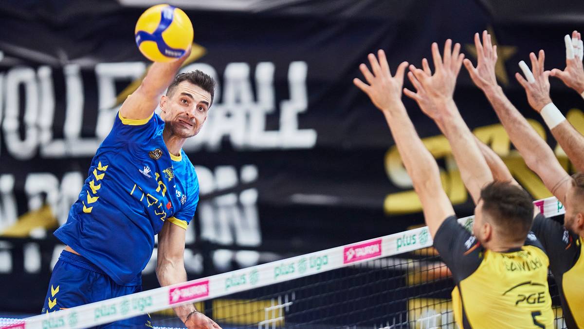 Siatkarz w niebieskiej koszulce z ż&oacute;łtymi akcentami wykonuje zagrywkę, podczas gdy przeciwnicy w ż&oacute;łtych strojach pr&oacute;bują go zablokować.