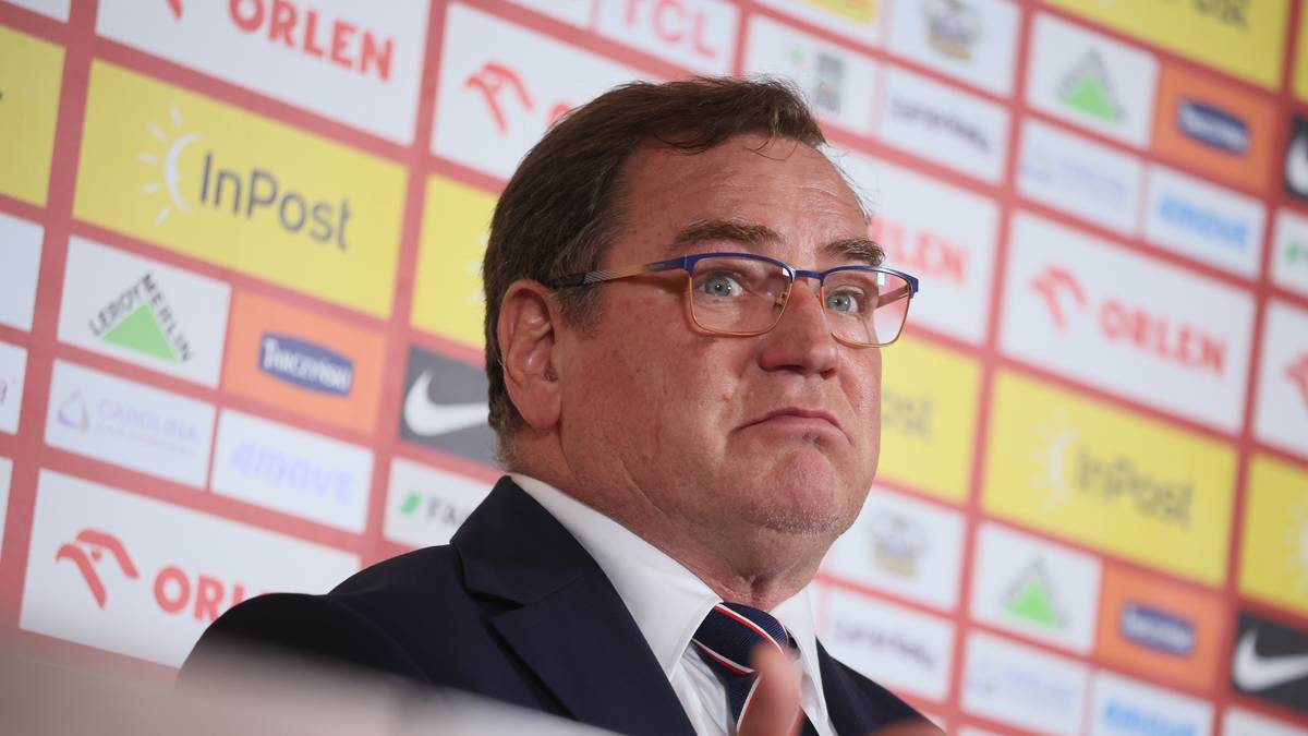 Portret mężczyzny w garniturze i okularach, stojącego przed ścianą z logotypami.