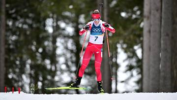 Biathlonistka w czerwonym stroju z numerem 7 na piersi, na nartach z kijami narciarskimi, na tle drzew i śniegu.