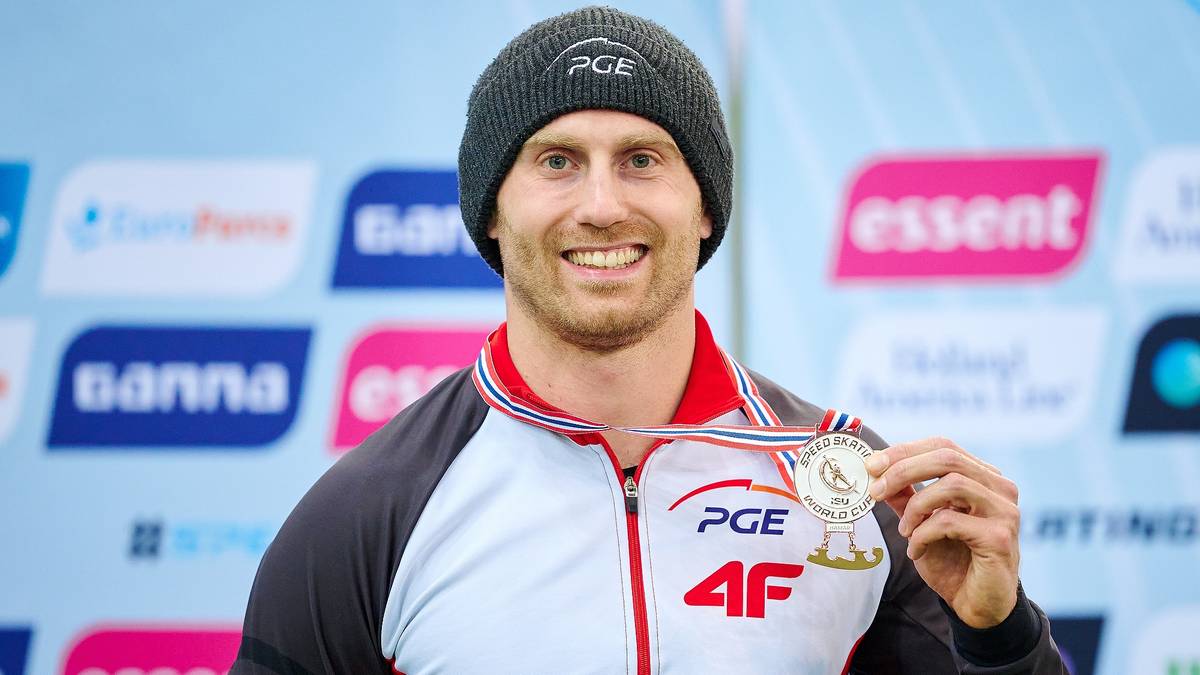 Uśmiechnięty mężczyzna w czapce i kurtce sportowej trzyma medal.