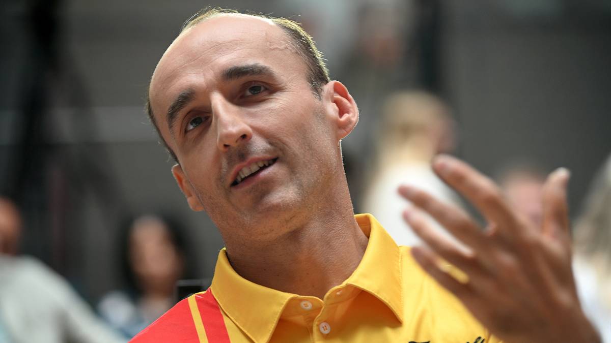 Robert Kubica w ż&oacute;łtej koszulce z czerwonymi pasami, gestykuluje prawą ręką.