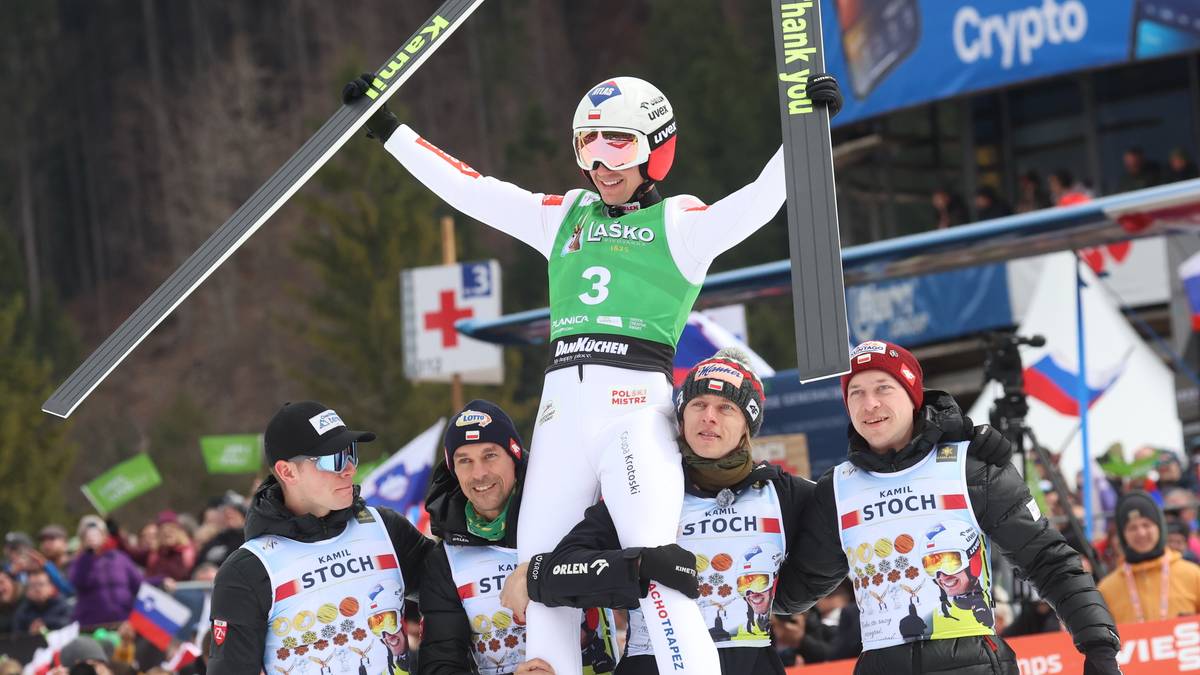 Kamil Stoch w zielonym plastronie z numerem 3, otoczony przez innych skoczków i trenerów, trzymający narty na tle tłumu kibiców.