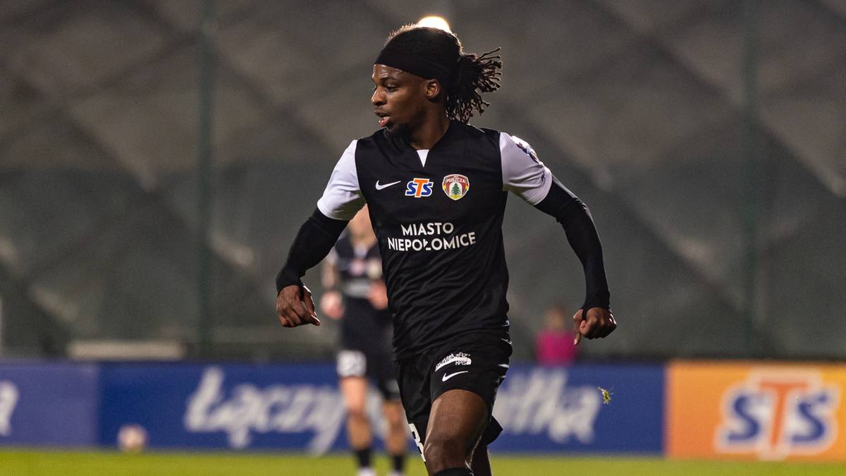 Piłkarz w czarno-białym stroju sportowym biegnie z piłką na boisku.