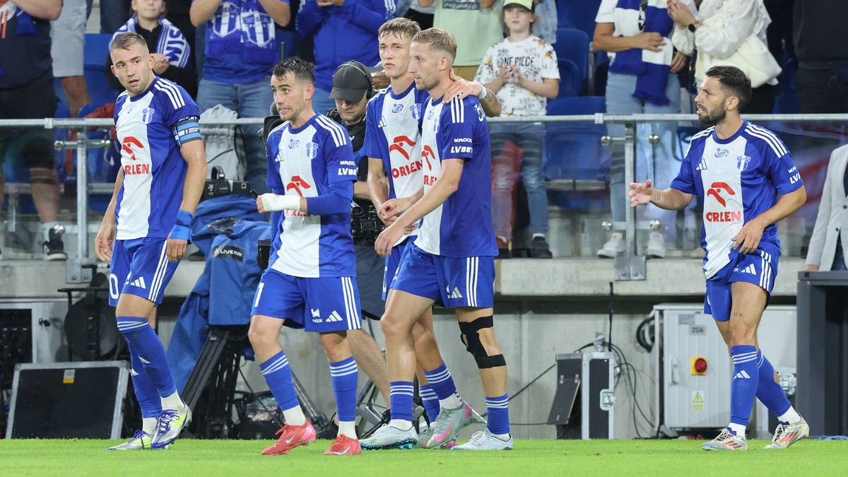 Grupa piłkarzy w niebiesko-białych strojach wychodzi na boisko, niektórzy patrzą w stronę widowni, inni w stronę kamery. W tle widać trybuny zapełnione kibicami.