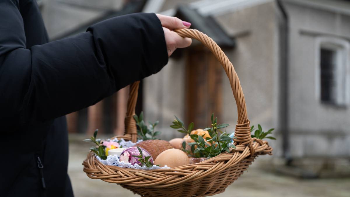 Dłoń trzymająca wiklinowy koszyczek z jajkami i zielonymi gałązkami, w tle budynek.