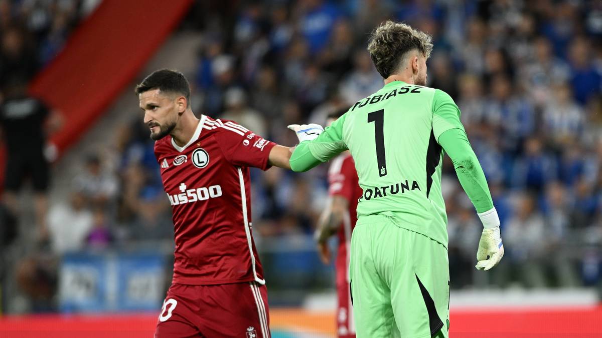 Dwóch piłkarzy, jeden w czerwonej koszulce z napisem "us500" i numerem 8 na plecach, drugi w zielonym stroju bramkarskim z numerem 1, stoją obok siebie na boisku piłkarskim.