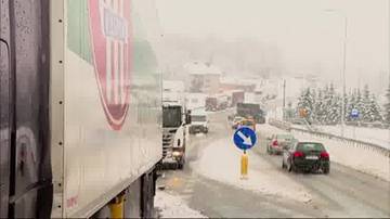 Ciężarówka i samochody osobowe stojące w korku na zaśnieżonej drodze. W tle widoczne budynki i drzewa pokryte śniegiem.