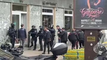 Policjanci w pełnym rynsztunku przed zdewastowanym budynkiem banku, z widocznym fragmentem samochodu na pierwszym planie.