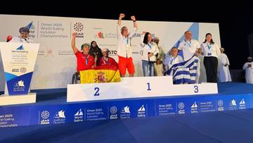 Trzech żeglarzy na podium, na środku zawodnik w białej koszulce podnosi ręce do g&oacute;ry, na pierwszym miejscu.
