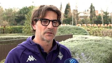 Paolo Vanoli w okularach i fioletowej bluzie z kapturem, m&oacute;wiący do mikrofonu na tle zieleni i budynk&oacute;w.