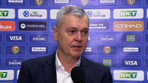 Ainars Bagatskis, trener koszyk&oacute;wki, w garniturze i koszuli, stoi przed ścianą z logo sponsor&oacute;w i napisami.