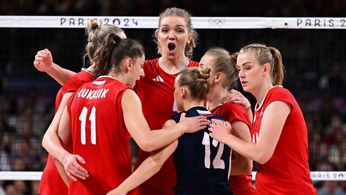 Siedem siatkarek w czerwonych strojach sportowych ze sobą rozmawia, częściowo się obejmując, w tle widać siatkę do siatkówki.