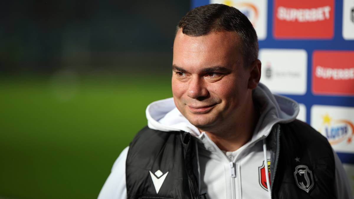 Mężczyzna w średnim wieku, ubrany w kamizelkę sportową i bluzę z kapturem, uśmiecha się lekko, stojąc na tle boiska piłkarskiego.