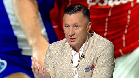 Mężczyzna w jasnej marynarce w kratę m&oacute;wi, gestykulując prawą ręką. Na jego piersi widoczna jest ozdobna chusteczka w kolorach czerwonym i niebieskim. W tle widoczni są inni mężczyźni w strojach sportowych.