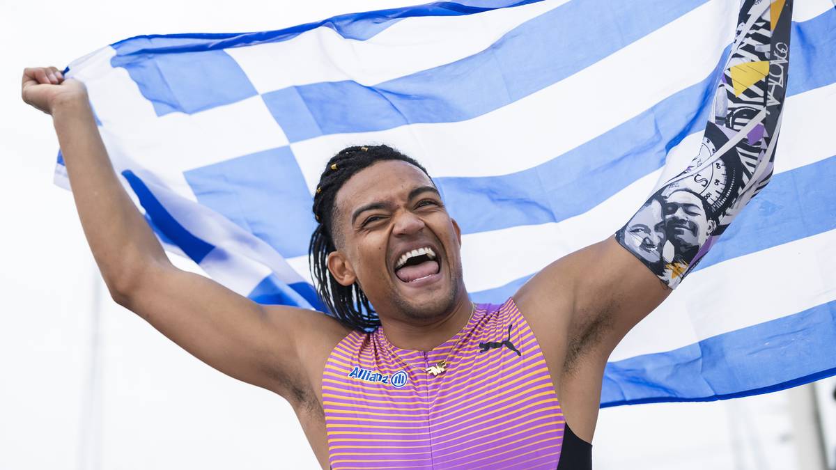 Szeroko uśmiechnięty Emmanuel Karalis trzyma grecką flagę nad głową.