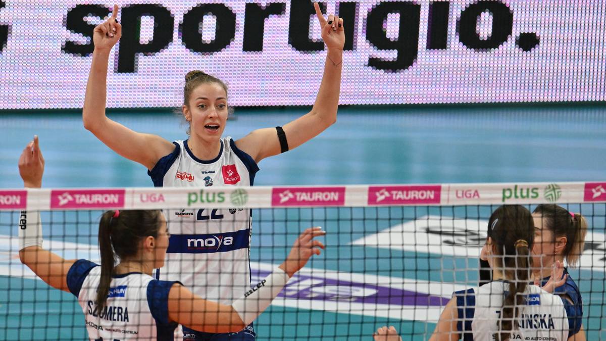 Siatkarka w białej koszulce z numerem 5 unosi ręce do góry w geście radości, patrząc w stronę widowni. Pozostałe zawodniczki stoją przy siatce, patrząc w stronę boiska.