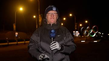 Mężczyzna w czapce i okularach z mikrofonem "Polsat Sport" w ręku, na tle nocnej iluminacji z pierścieniami olimpijskimi.