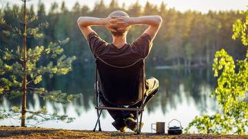 Mężczyzna siedzi na krześle w plenerze, patrząc na jezioro i las.
