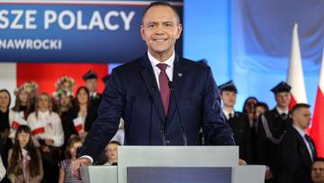 Uśmiechnięty mężczyzna w garniturze, stojący za m&oacute;wnicą z napisem "POLSKI SEJM 0%"