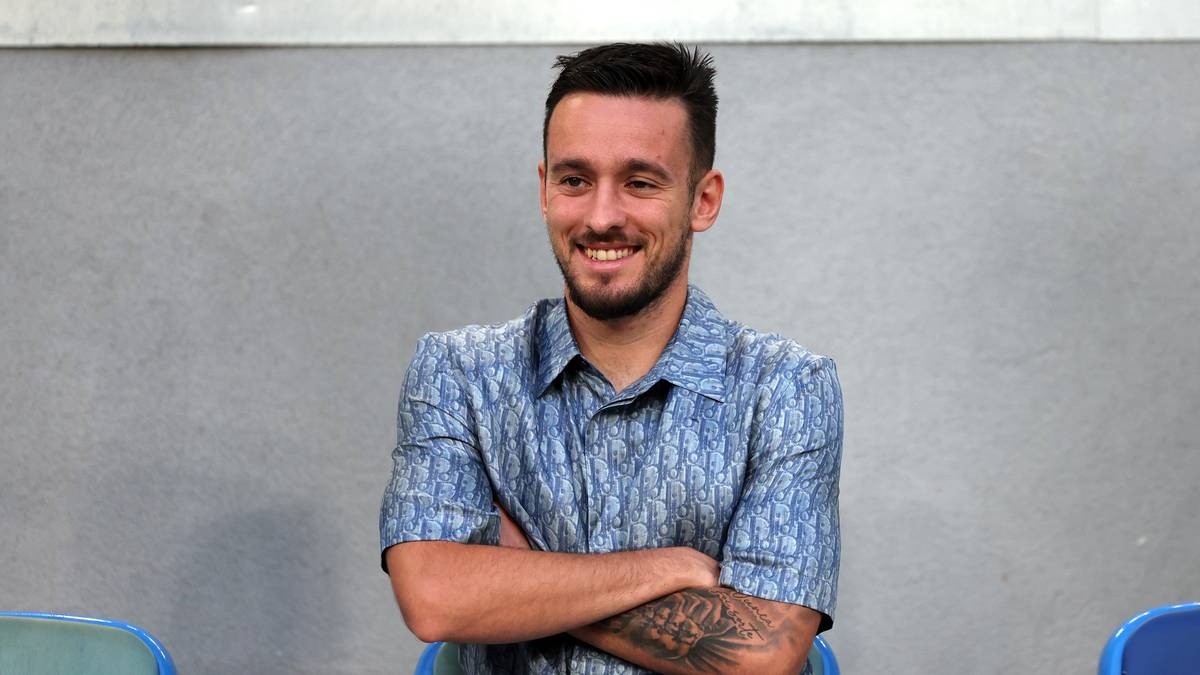 Uśmiechnięty mężczyzna w niebieskiej koszuli w wzory, z rękami skrzyżowanymi na piersi.