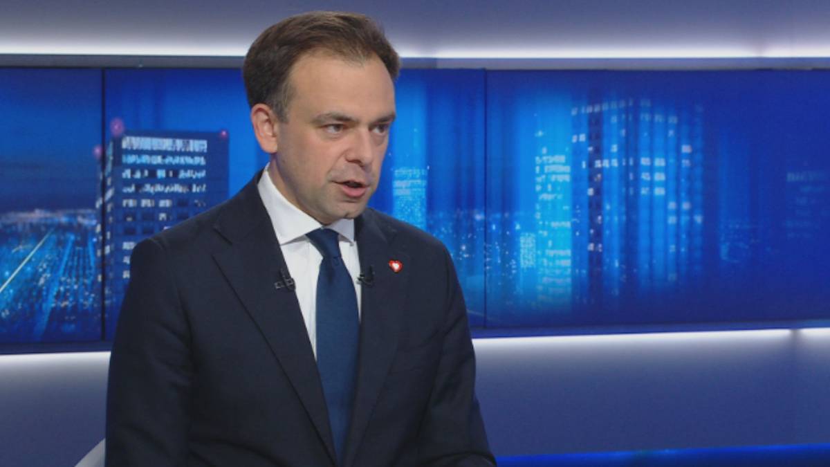 Mężczyzna w garniturze i krawacie przemawia, w tle widać ekran z widokiem nocnego miasta.