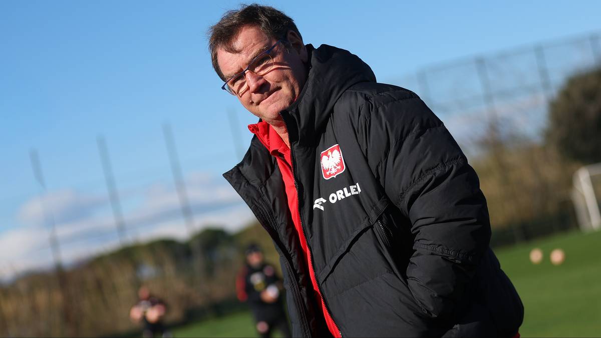 Mężczyzna w kurtce z godłem Polski uśmiecha się na tle boiska piłkarskiego.