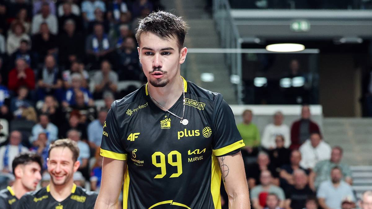 Siatkarz z numerem 99 na koszulce prowadzi drużynę na boisko, w tle kibice na trybunach.