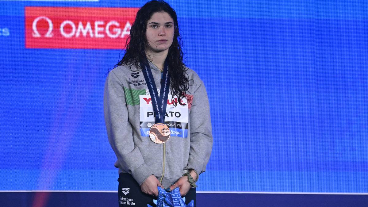 Młoda kobieta w szarej bluzie z kapturem i medalem na szyi stoi na podium, patrząc przed siebie ze smutną miną.