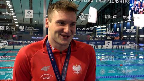 Pływak w czerwonej bluzie z medalem na szyi uśmiecha się na tle basenu.