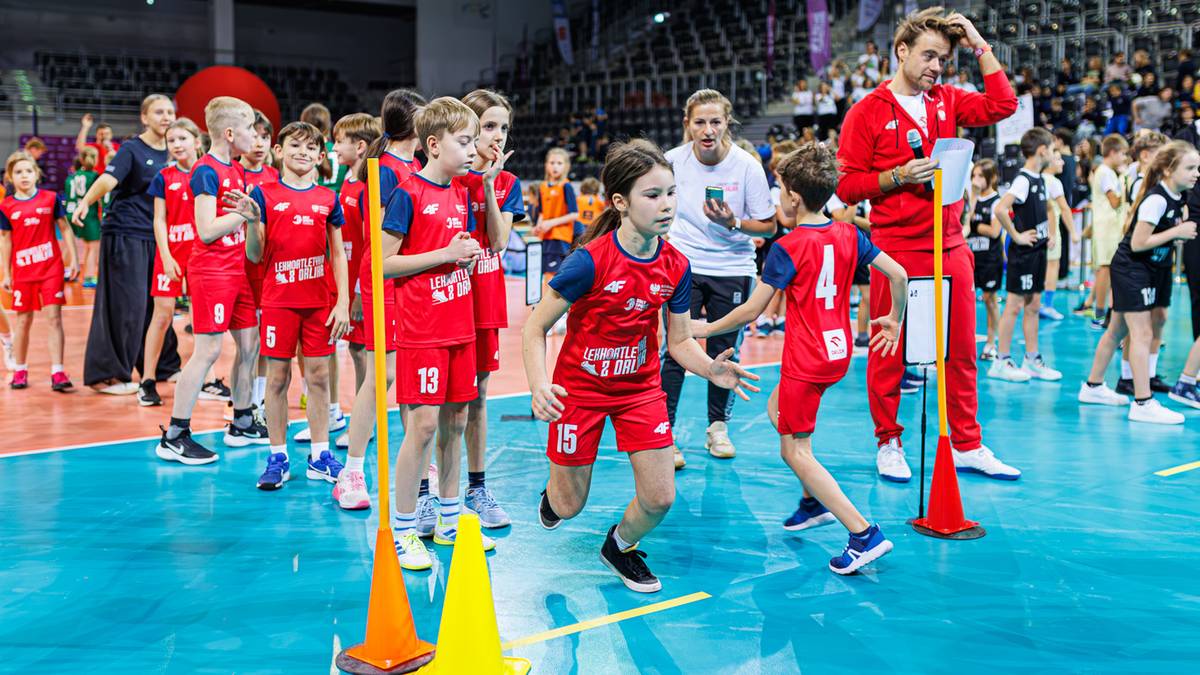 Grupa dzieci w czerwonych strojach sportowych uczestniczy w zawodach lekkoatletycznych, z trenerami i ambasadorem programu.