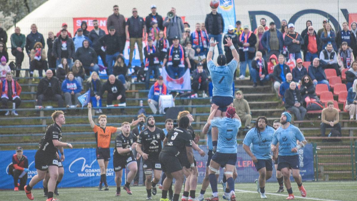 Zawodnicy drużyny rugby w jasnoniebieskich strojach wykonują podnoszenie zawodnika do chwytu piłki z autu, podczas gdy na trybunach zasiadają kibice.