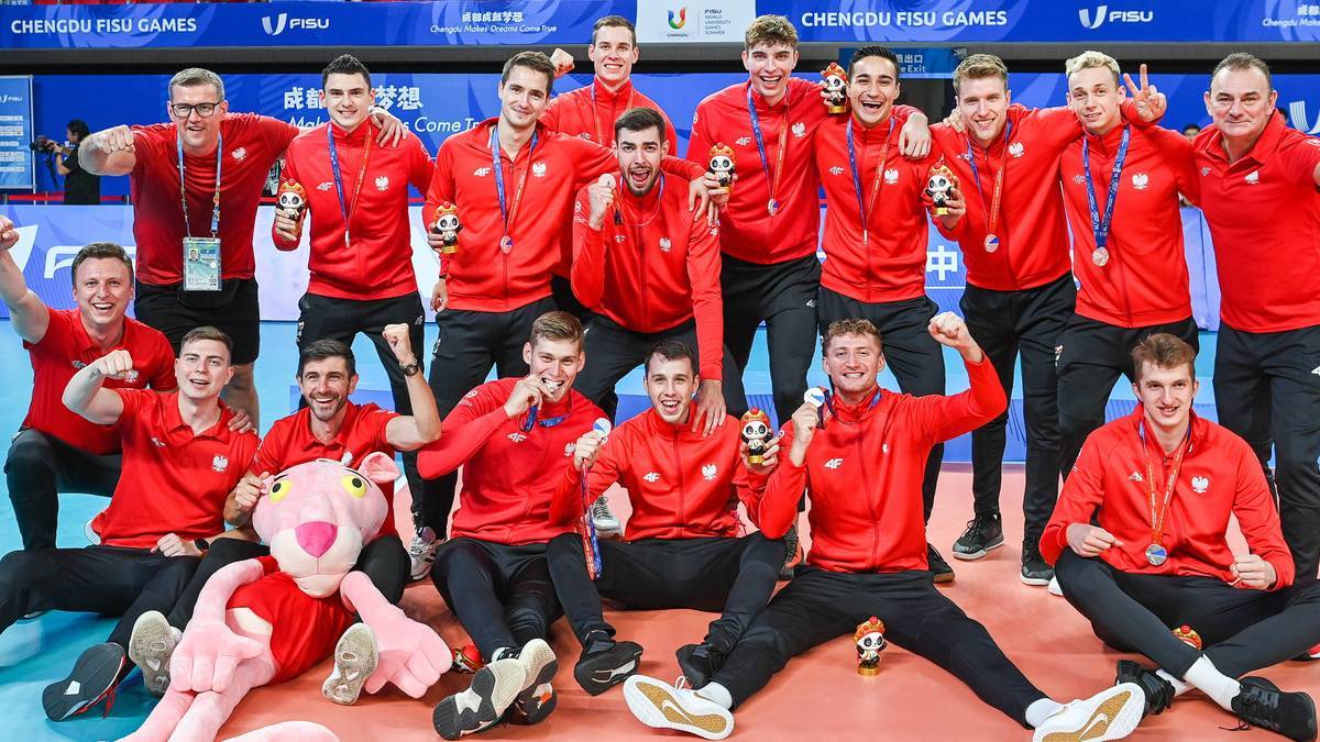 Grupa młodych mężczyzn w czerwonych koszulkach i spodenkach pozuje do zdjęcia z pucharem i maskotką.