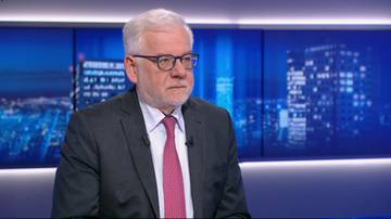 Starszy mężczyzna w okularach i garniturze, siedzący na tle ekranu z widokiem na nocne miasto.