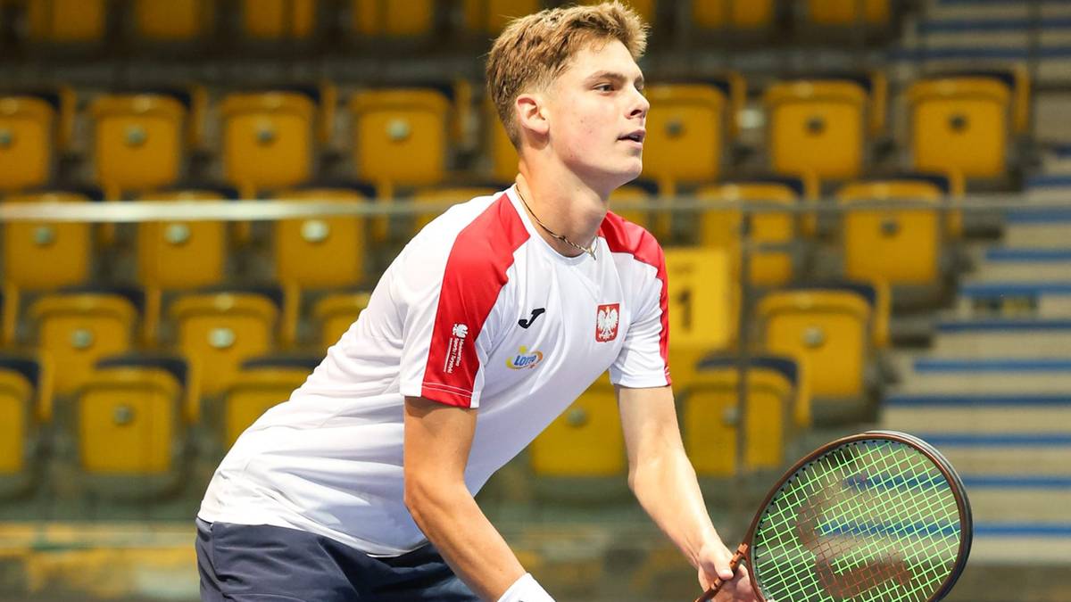 Młody mężczyzna, tenisista, w białej koszulce z czerwonymi elementami, gra w tenisa. Jest na korcie tenisowym z żółtymi trybunami w tle.