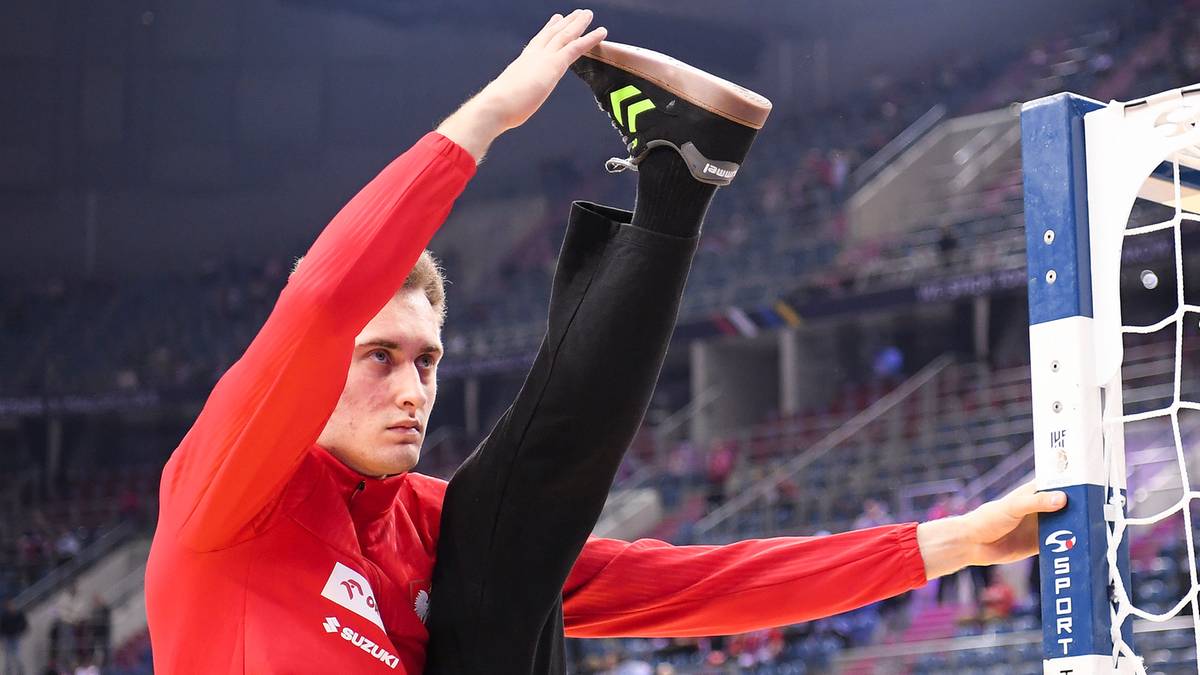 Piłkarz ręczny w czerwonej koszulce rozciąga nogę, opierając stopę na poprzeczce bramki na hali sportowej.