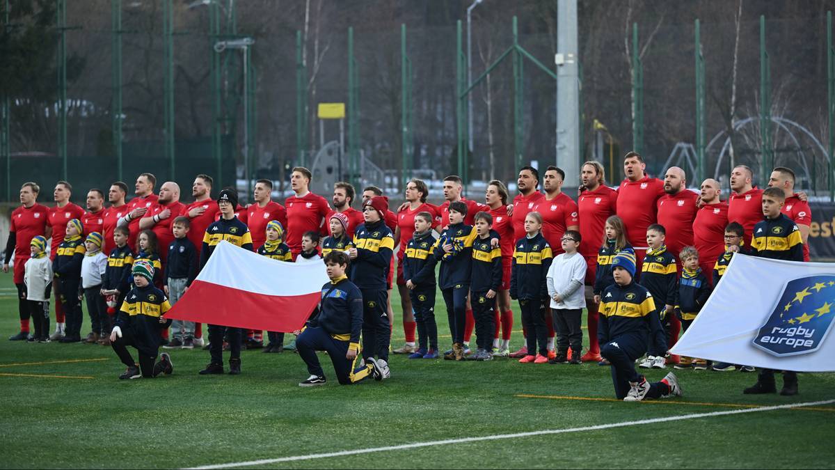 Reprezentacja Polski w rugby stoi na boisku z biało-czerwoną flagą.