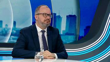 Mężczyzna w okularach i garniturze siedzi w studiu telewizyjnym, na stole stoi szklanka z wodą. W tle widoczna jest grafika miejska.