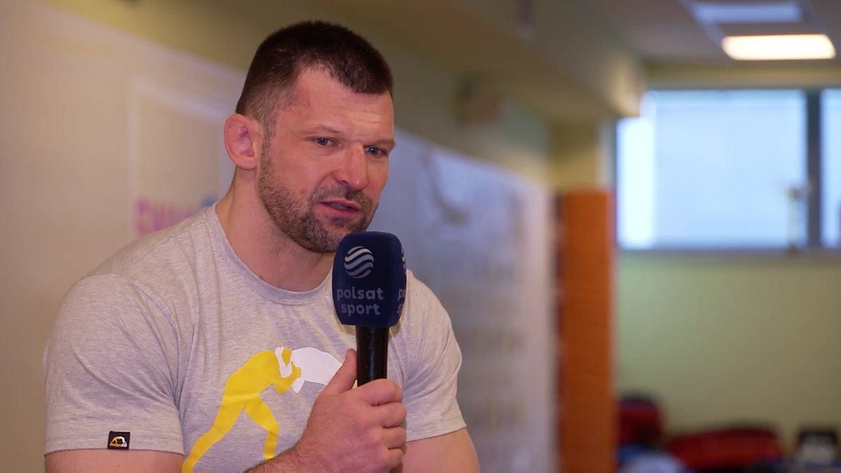 Szymon Kołecki w szarym t-shircie z grafiką przedstawiającą sztangistę, trzymający mikrofon Polsat Sport podczas wywiadu.